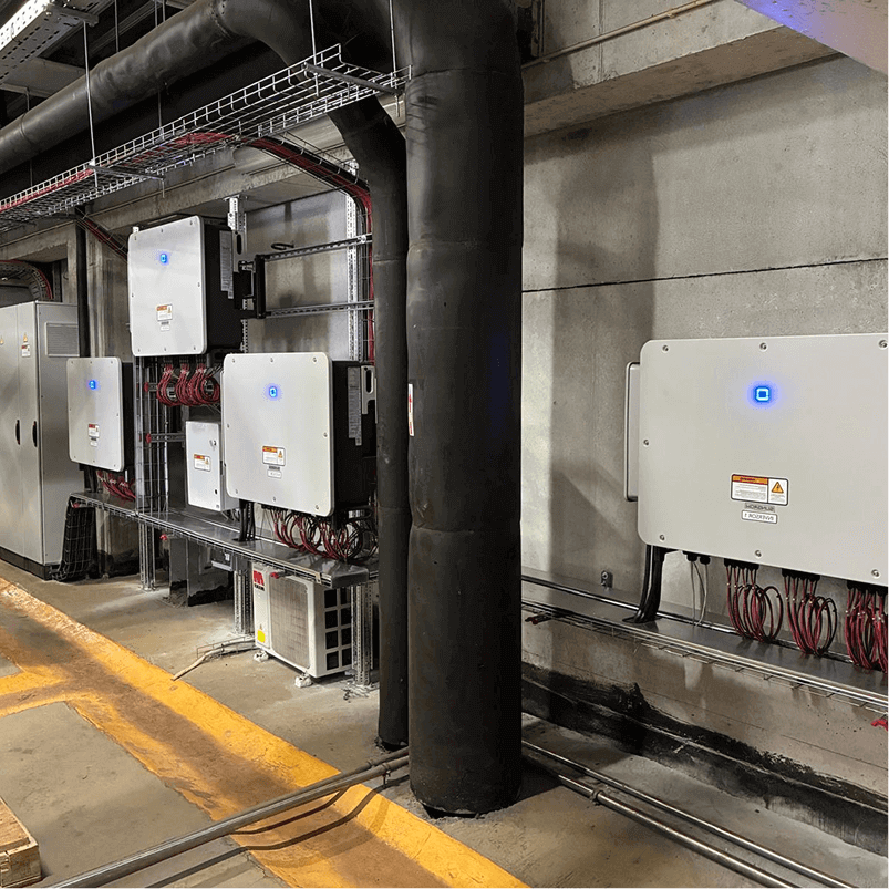 Row of electrical inverters mounted on a concrete wall with visible cables and pipes.